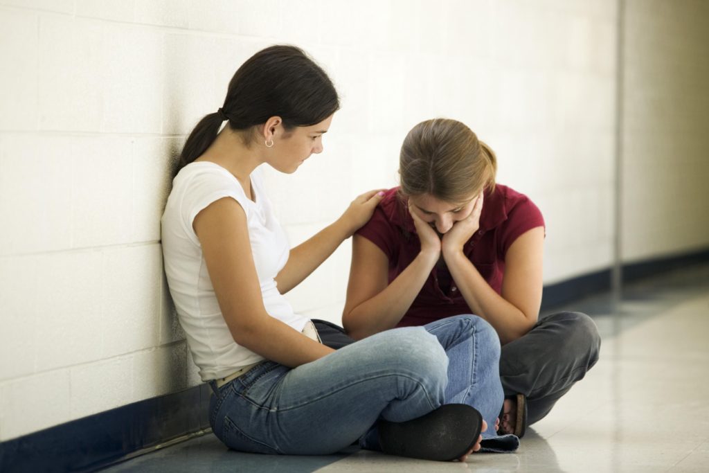 two teens in a hall, one consoling the other who appears depressed.